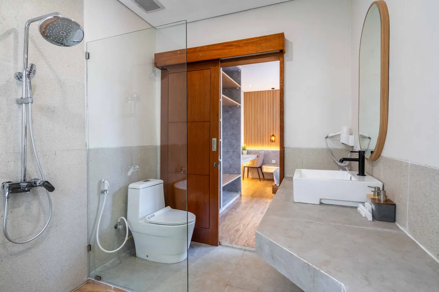 Modern minimalist bathroom featuring a freestanding white bathtub, sleek vanity sink with black faucet, round wooden mirror, and tropical plant-filled window. - Bali Villas