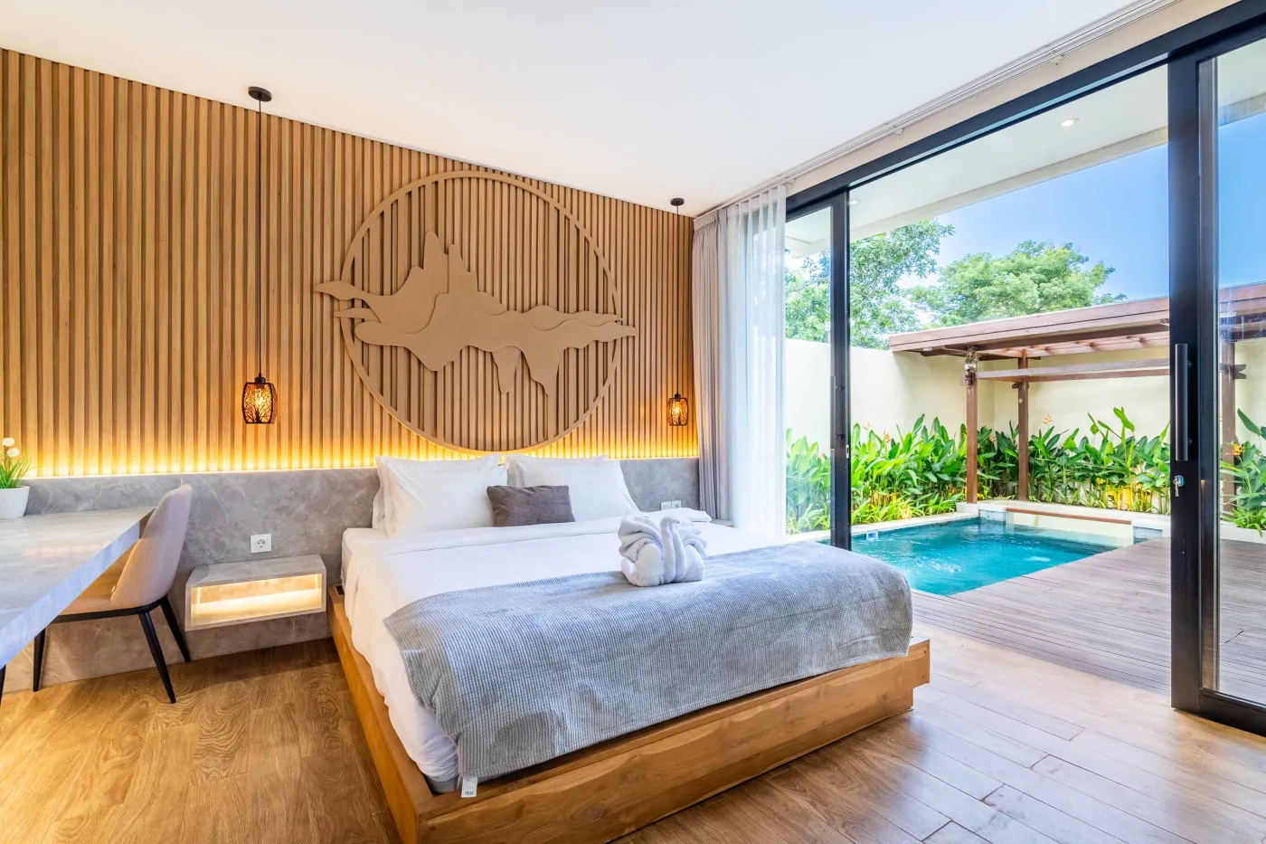 Modern minimalist bathroom featuring a freestanding white bathtub, sleek vanity sink with black faucet, round wooden mirror, and tropical plant-filled window. - Bali Villas