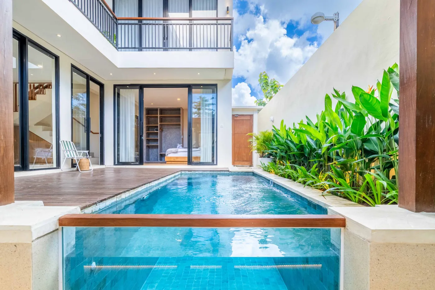 Modern minimalist bathroom featuring a freestanding white bathtub, sleek vanity sink with black faucet, round wooden mirror, and tropical plant-filled window. - Bali Villas