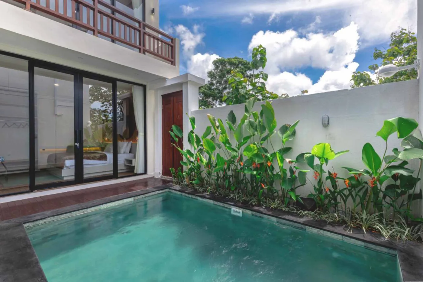 Serene tropical villa pool with clear turquoise water, surrounded by lush plants and modern glass-walled house under a blue sky. - Bali Villas