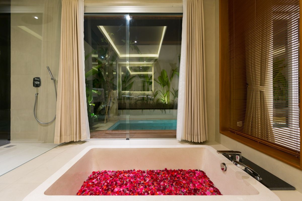 Luxurious Balinese villa terrace at night: wooden table with red petals, white armchairs, tropical banana plants, and sliding glass doors to bedroom. - Bali Villas