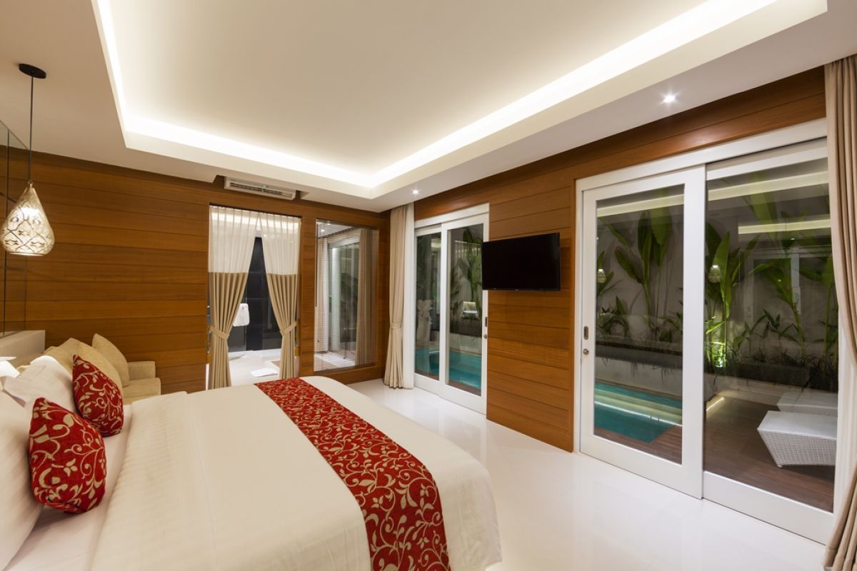 Luxurious Balinese villa terrace at night: wooden table with red petals, white armchairs, tropical banana plants, and sliding glass doors to bedroom. - Bali Villas