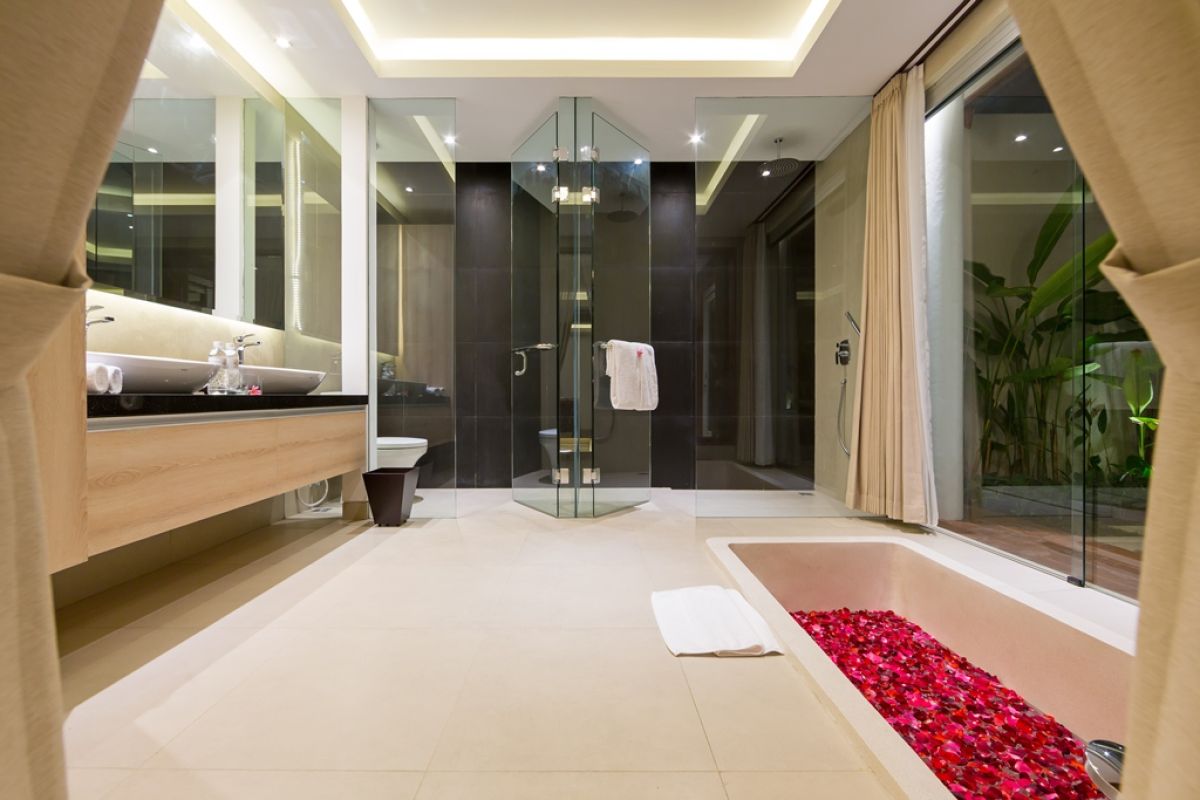 Luxurious Balinese villa terrace at night: wooden table with red petals, white armchairs, tropical banana plants, and sliding glass doors to bedroom. - Bali Villas