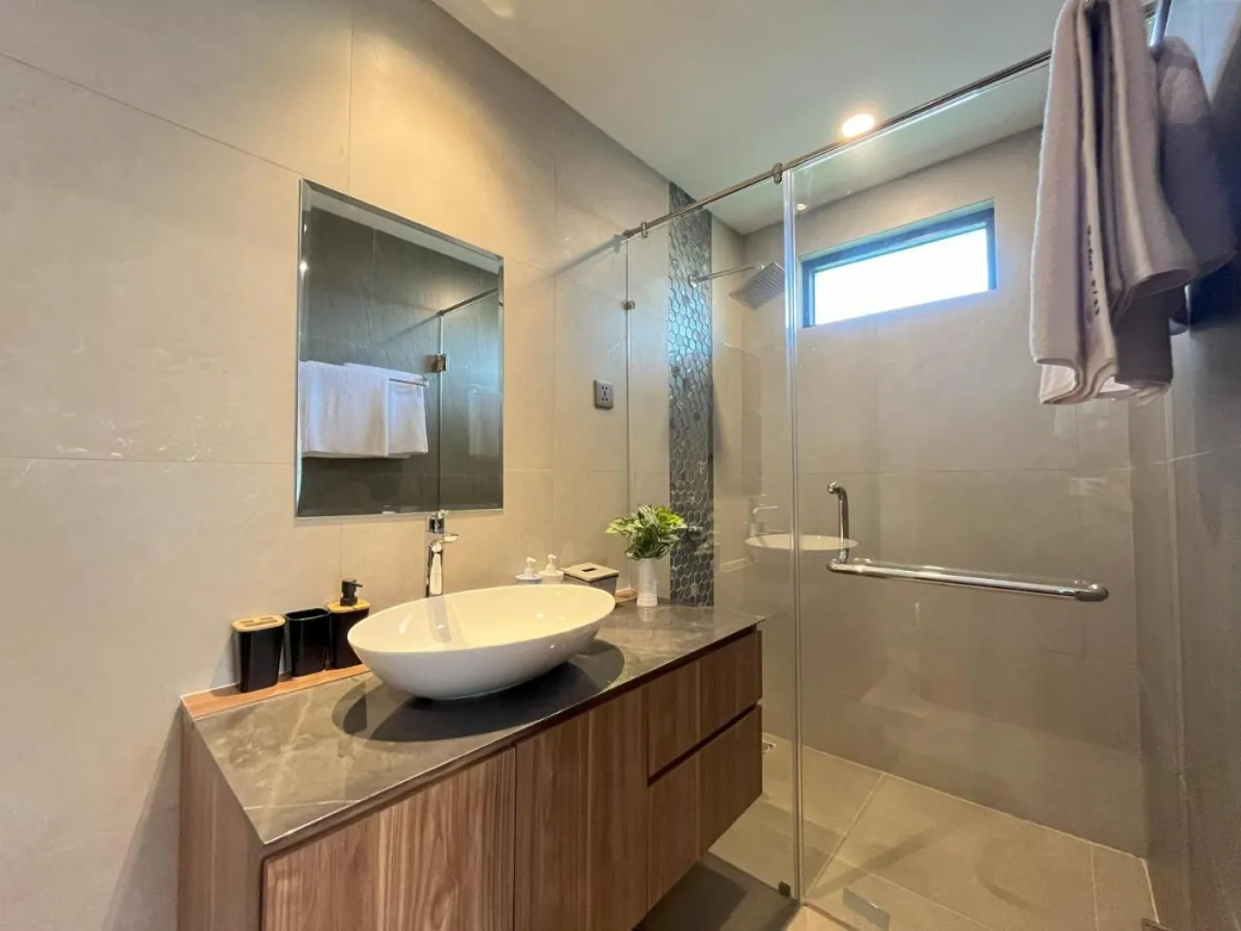 Modern gray-tiled bathroom with wooden vanity, white vessel sink, glass shower, and hanging towels. - Bali Villas
