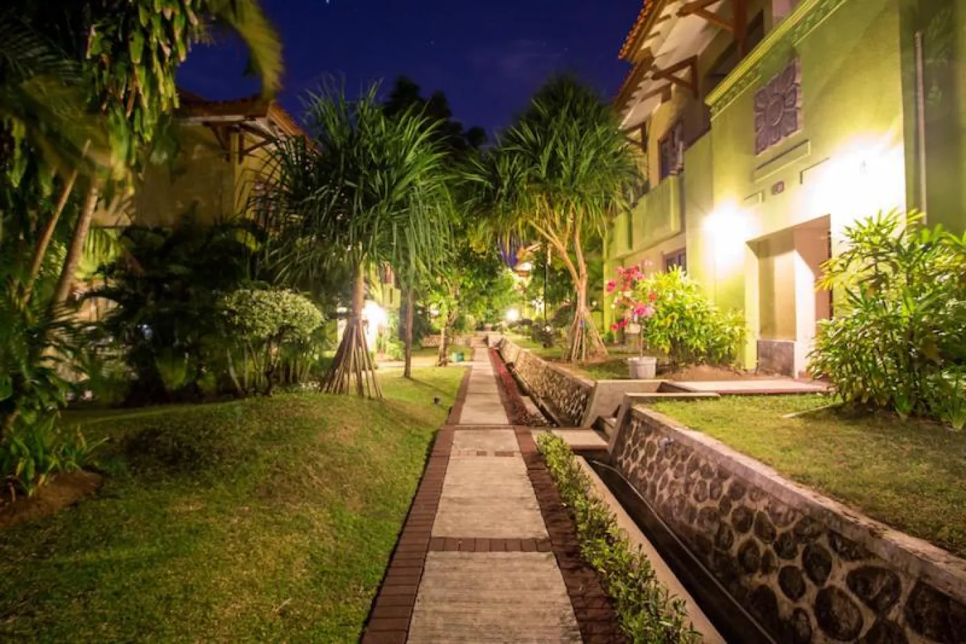 Illuminated nighttime pathway through lush tropical gardens with palm trees, green resort buildings, and stone water channel. - Bali Villas