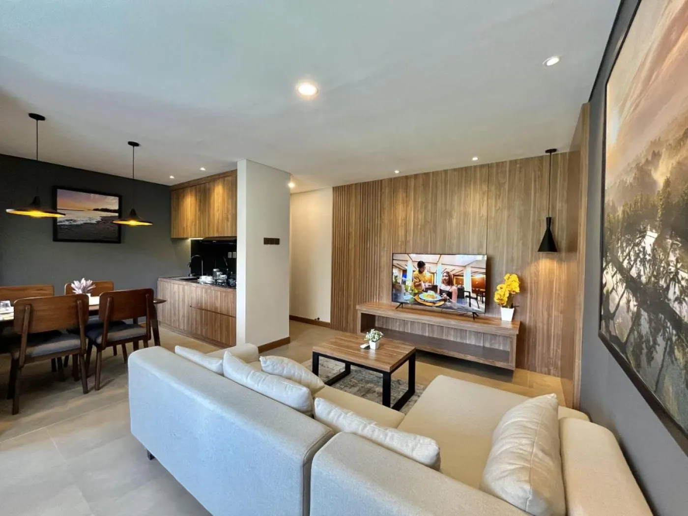 Modern open-plan living room with wooden paneling, white L-shaped sofa, dining table, kitchen cabinets, and large TV in a neutral interior. - Bali Villas