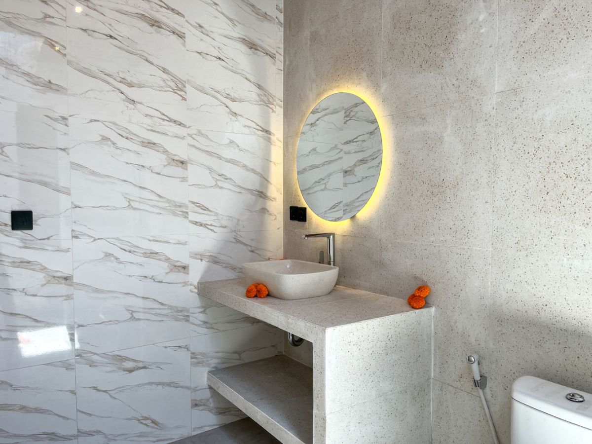 Modern Balinese kitchen with warm teak cabinets, white marble counters, stainless steel appliances, and open archway to outdoor patio with plants and seating. - Bali Villas