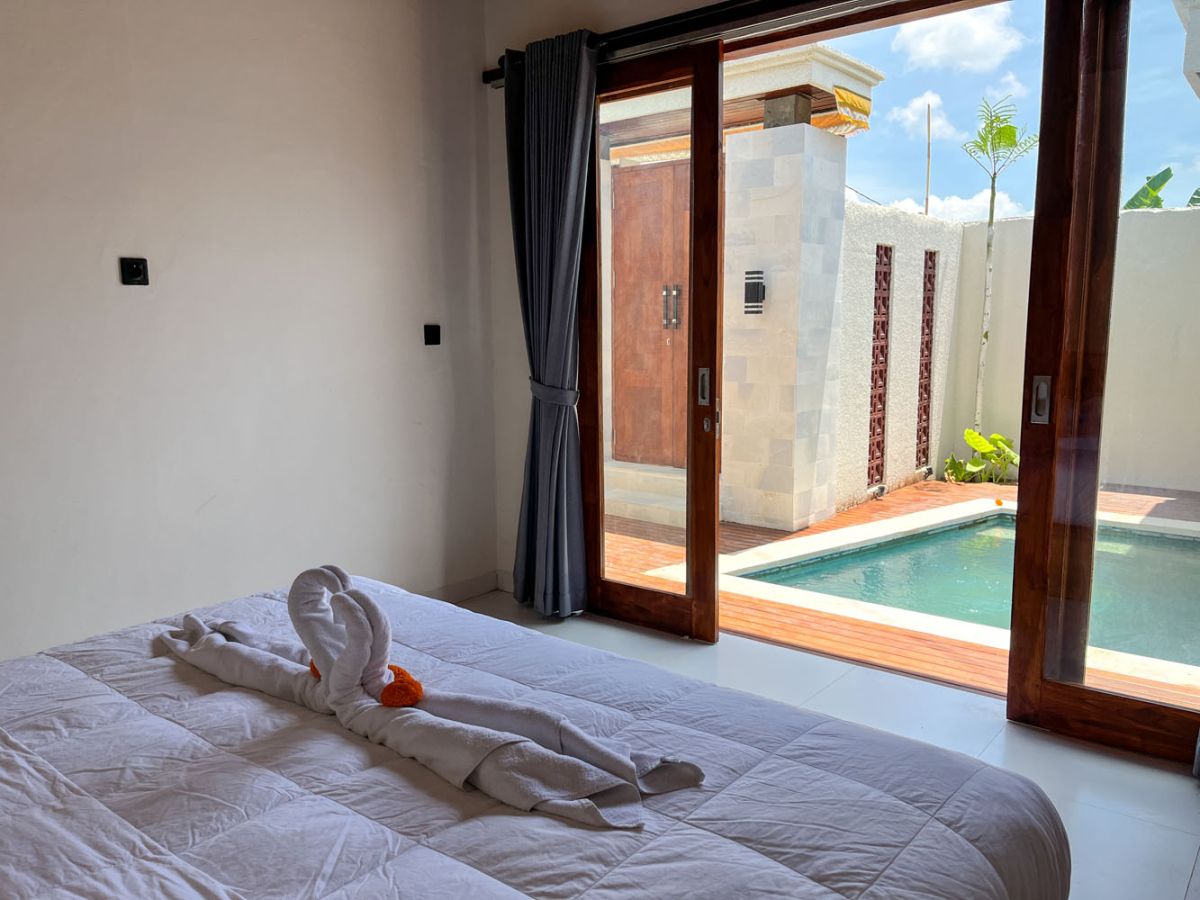 Modern Balinese kitchen with warm teak cabinets, white marble counters, stainless steel appliances, and open archway to outdoor patio with plants and seating. - Bali Villas