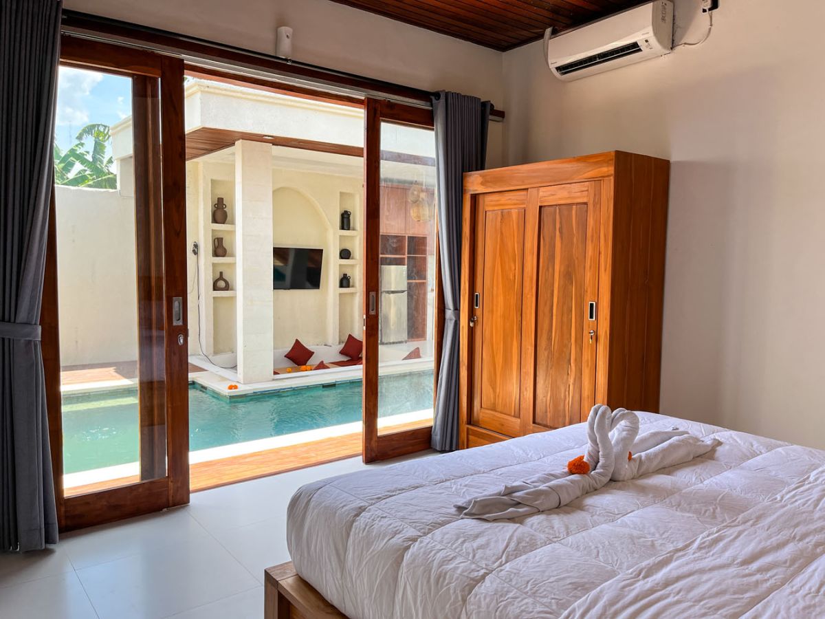 Modern Balinese kitchen with warm teak cabinets, white marble counters, stainless steel appliances, and open archway to outdoor patio with plants and seating. - Bali Villas