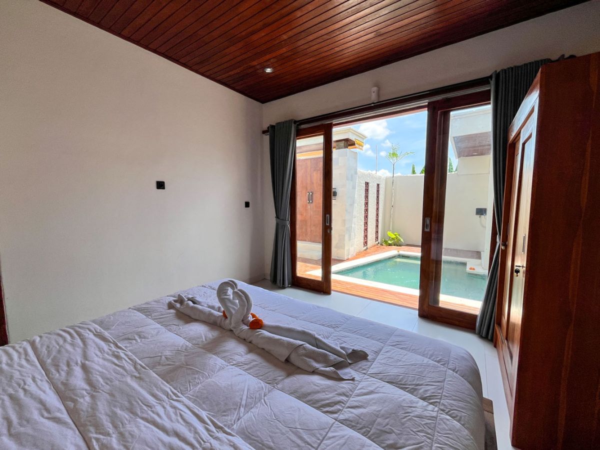 Modern Balinese kitchen with warm teak cabinets, white marble counters, stainless steel appliances, and open archway to outdoor patio with plants and seating. - Bali Villas