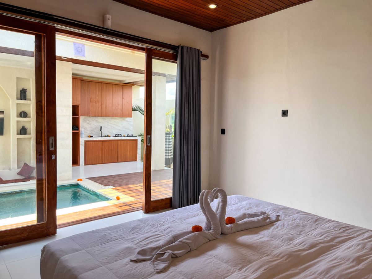 Modern Balinese kitchen with warm teak cabinets, white marble counters, stainless steel appliances, and open archway to outdoor patio with plants and seating. - Bali Villas