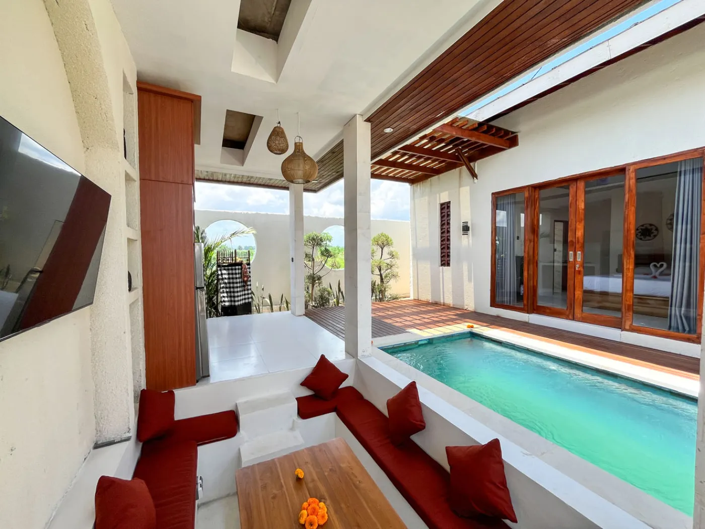 Serene open-air Bali villa lounge with red cushions, wooden table of tropical flowers, and turquoise private pool amid lush plants. - Bali Villas