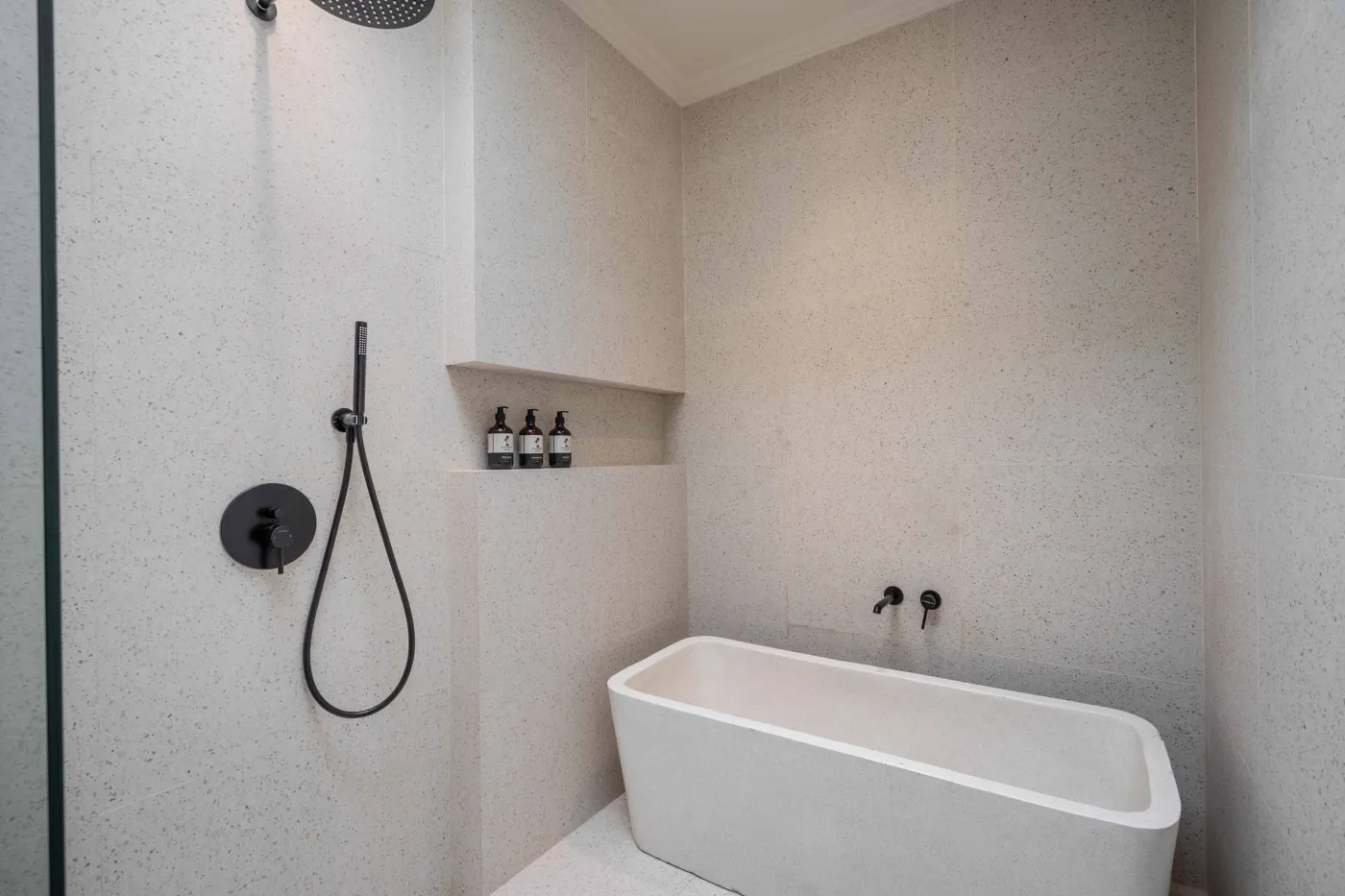 Modern minimalist bathroom with white freestanding bathtub, black rainfall showerhead, and sleek wall niche holding toiletries. - Bali Villas