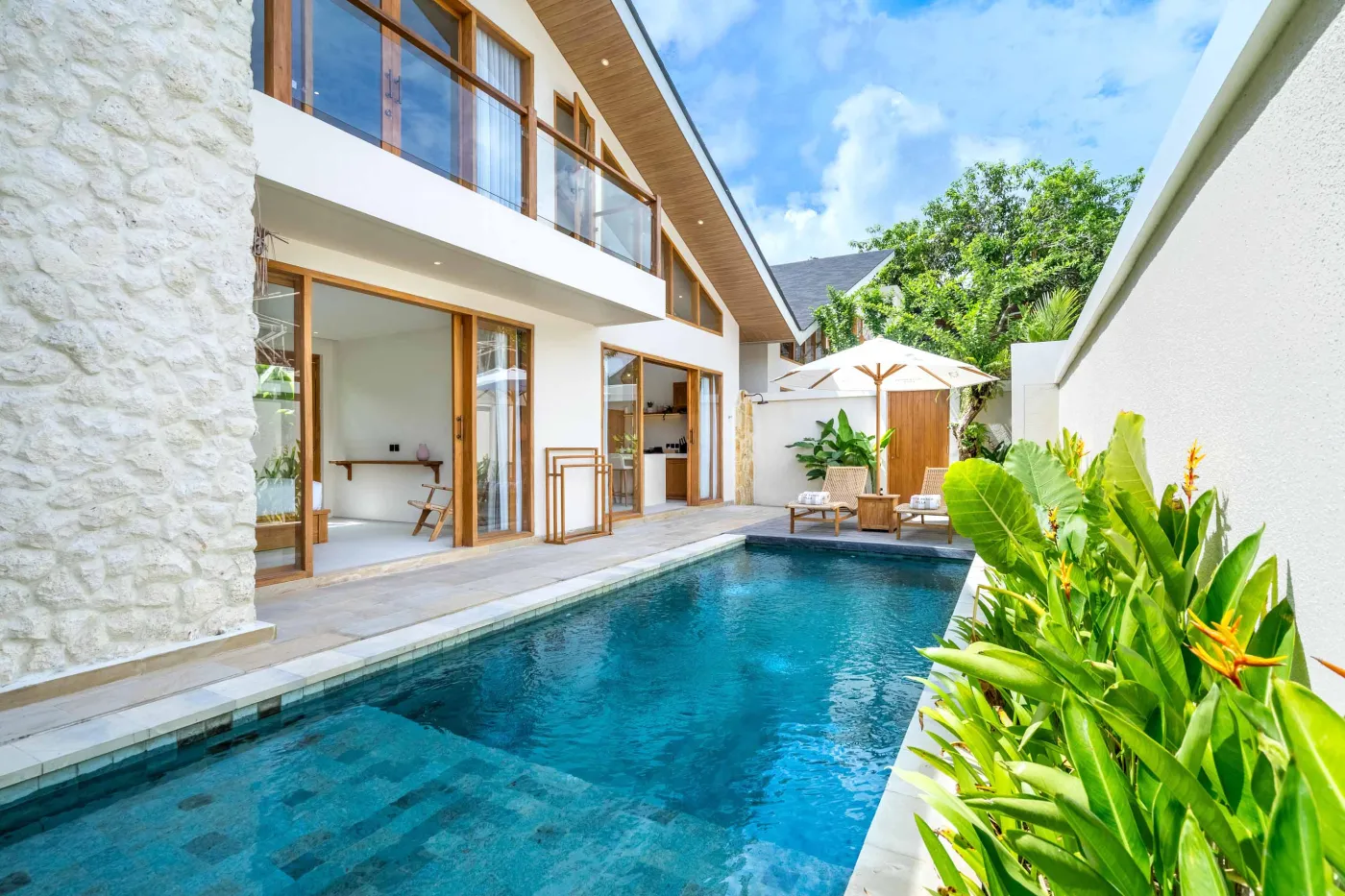 Modern tropical villa with infinity pool, sliding glass doors, and lush garden under a blue sky. - Bali Villas