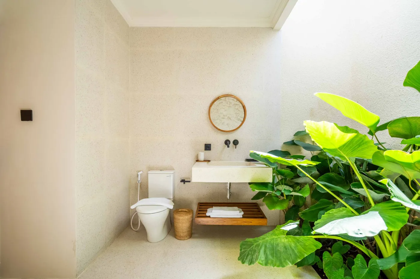 Luxurious tropical bathroom with white freestanding bathtub, rainfall shower, lush green plants, and textured stone walls. - Bali Villas