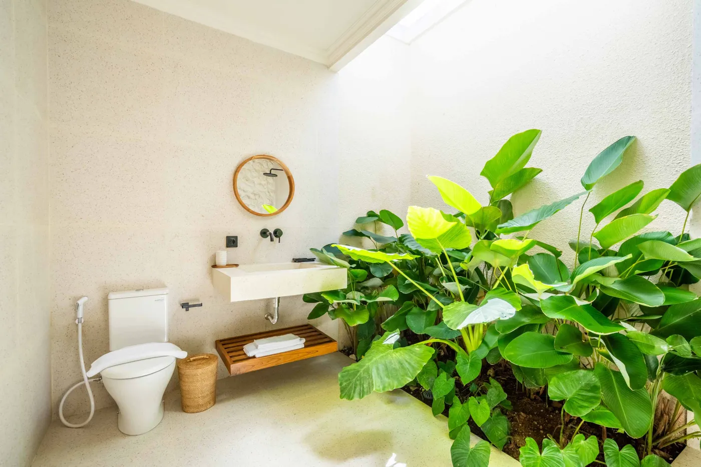 Luxurious tropical bathroom with white freestanding bathtub, rainfall shower, lush green plants, and textured stone walls. - Bali Villas