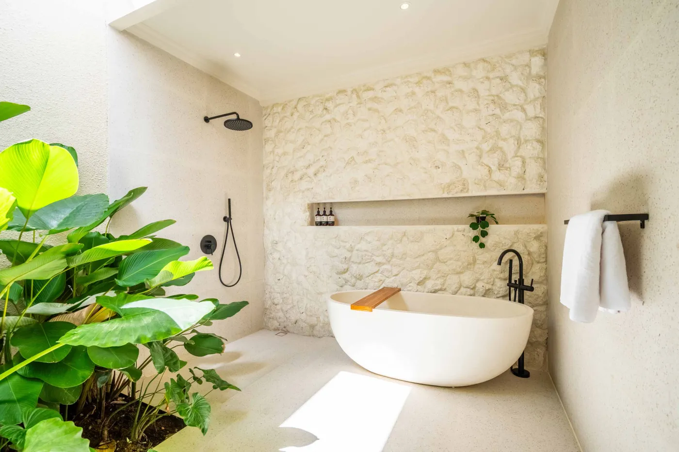 Luxurious tropical bathroom with white freestanding bathtub, rainfall shower, lush green plants, and textured stone walls. - Bali Villas