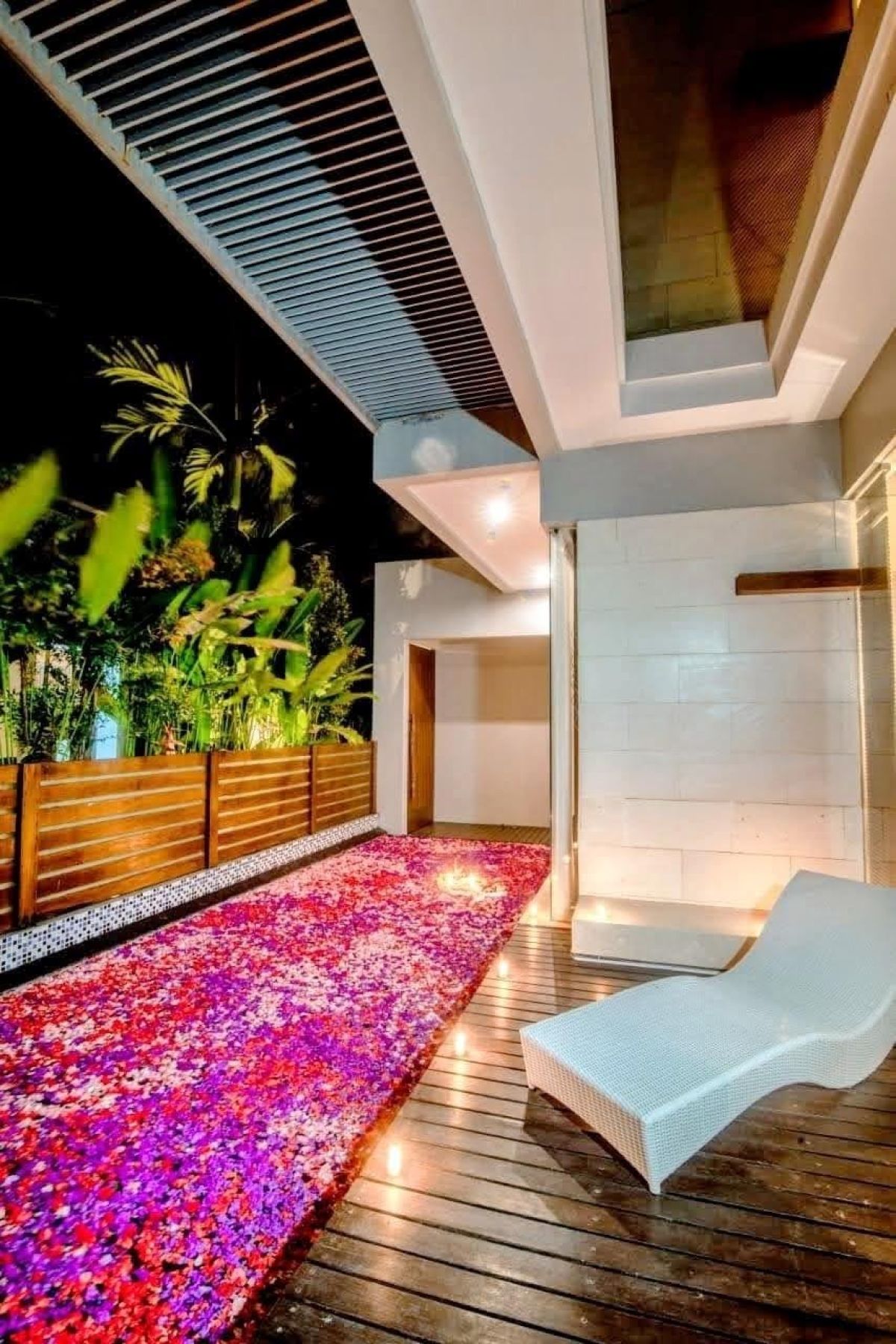 Modern Indonesian house exterior with white walls, orange accents, stone and wood features, parked scooter, and tropical plants on a sunny street. - Bali Villas