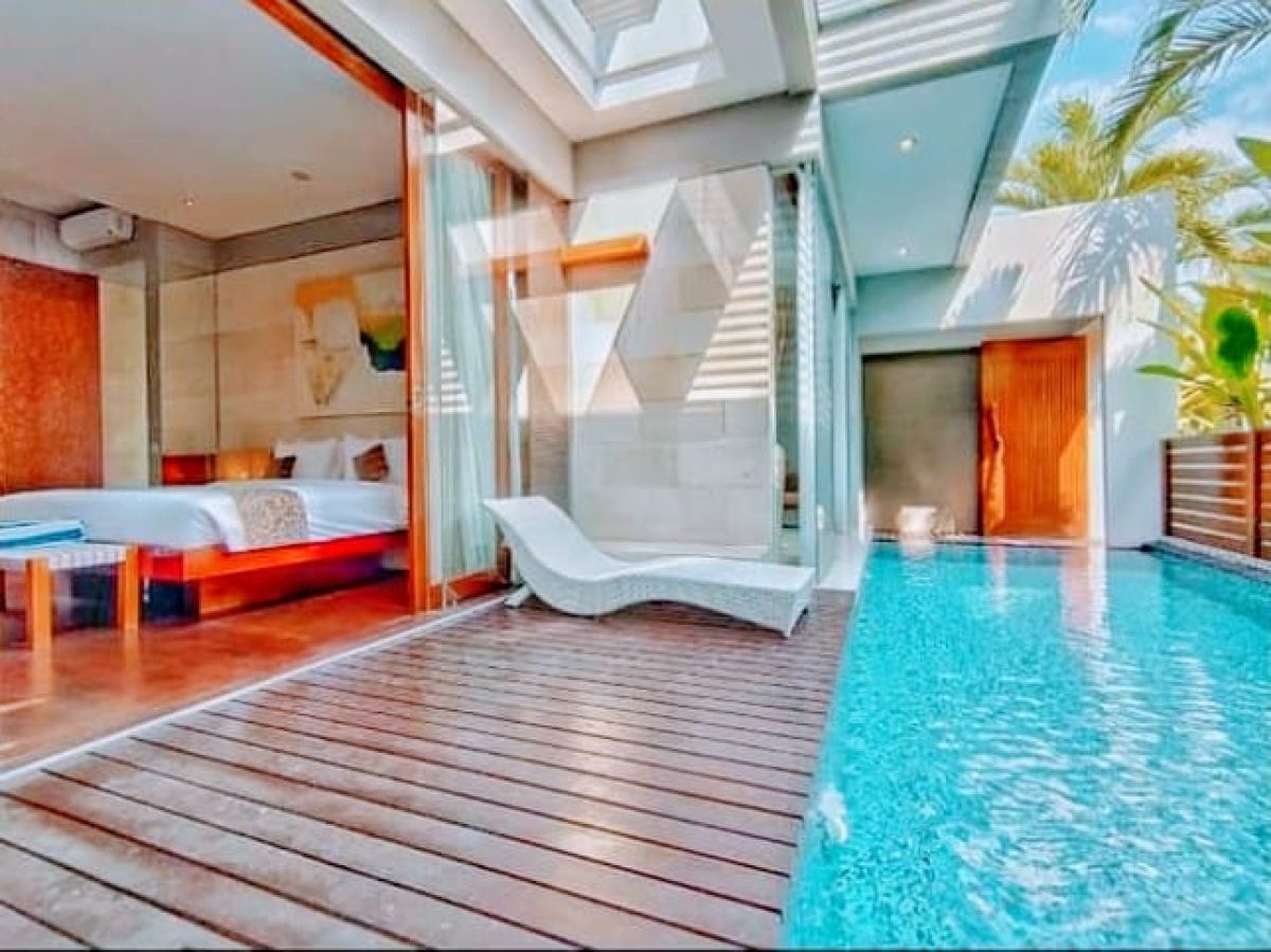 Modern Indonesian house exterior with white walls, orange accents, stone and wood features, parked scooter, and tropical plants on a sunny street. - Bali Villas