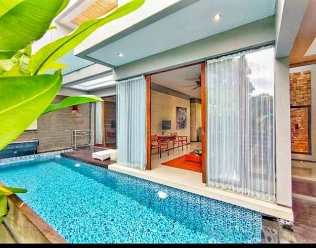 Modern Indonesian house exterior with white walls, orange accents, stone and wood features, parked scooter, and tropical plants on a sunny street. - Bali Villas