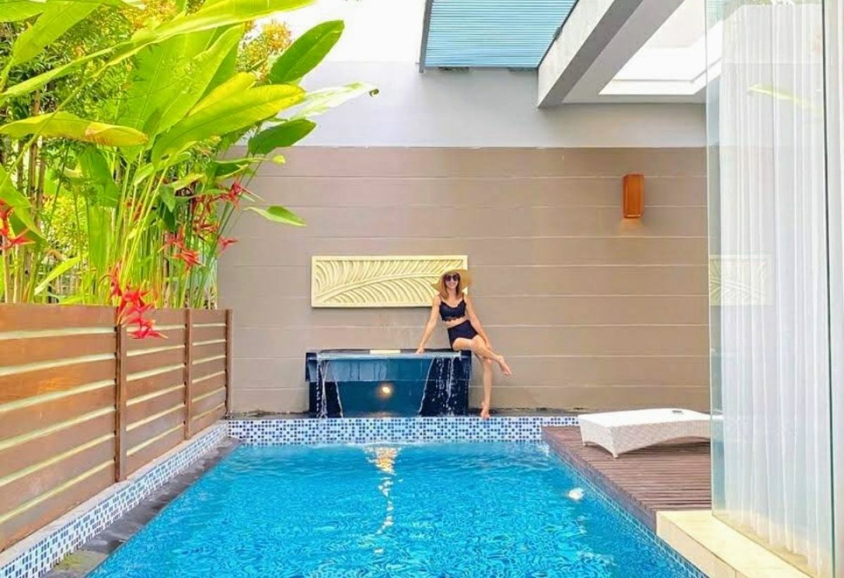Modern Indonesian house exterior with white walls, orange accents, stone and wood features, parked scooter, and tropical plants on a sunny street. - Bali Villas