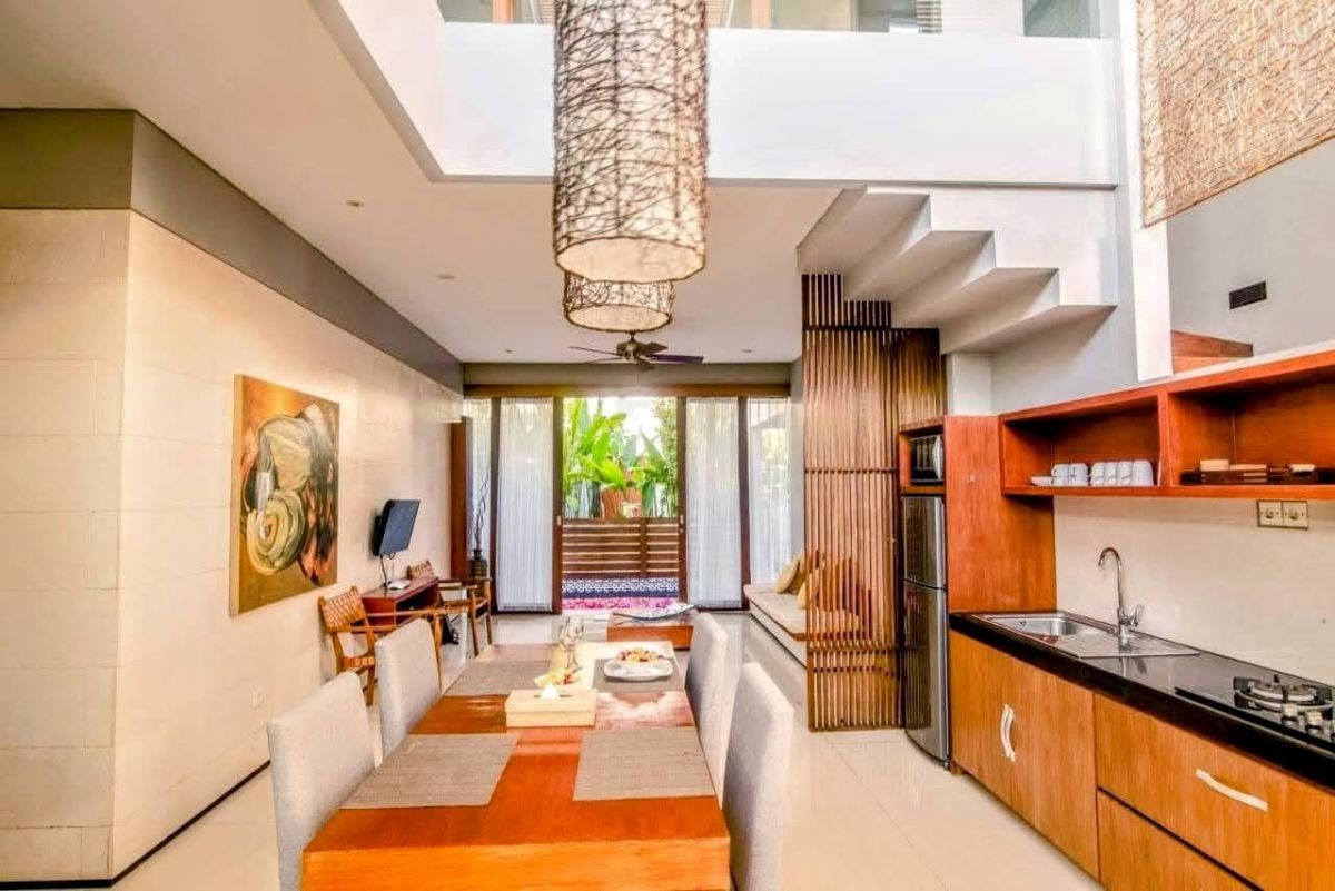 Modern Indonesian house exterior with white walls, orange accents, stone and wood features, parked scooter, and tropical plants on a sunny street. - Bali Villas