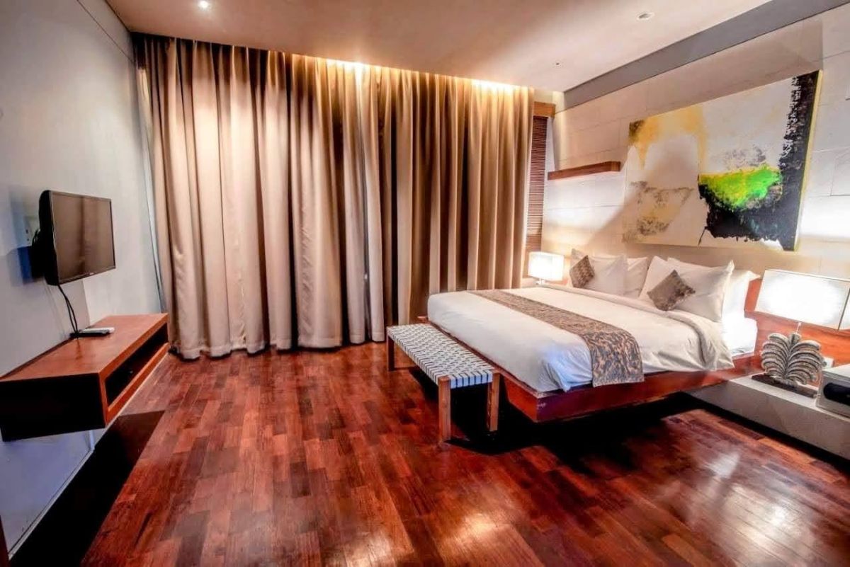 Modern luxury bedroom with king-sized bed, abstract yellow-green wall art, bedside lamps, beige curtains, and woven bench on wooden floor. - Bali Villas