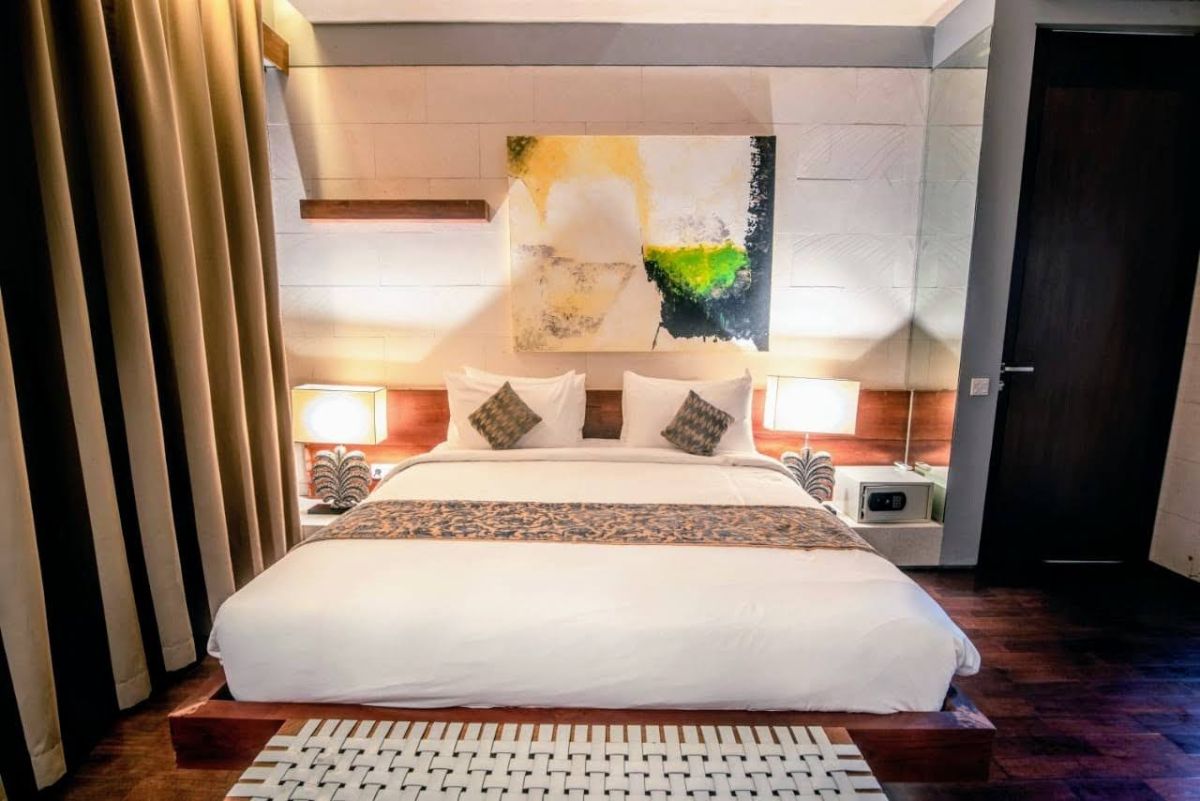 Modern luxury bedroom with king-sized bed, abstract yellow-green wall art, bedside lamps, beige curtains, and woven bench on wooden floor. - Bali Villas