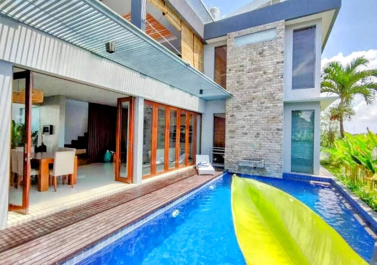 Modern tropical villa with infinity pool, wooden deck, sliding glass doors, and lush palm surroundings. - Bali Villas