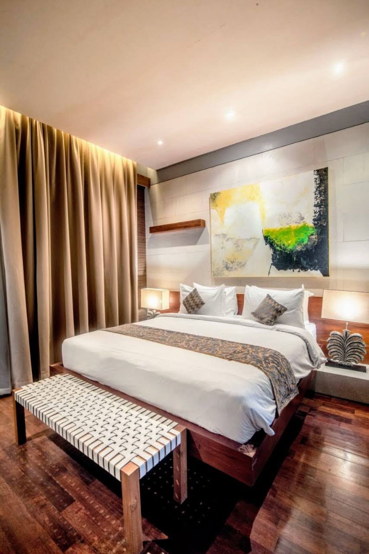 Modern luxury bedroom with king-sized bed, abstract yellow-green wall art, bedside lamps, beige curtains, and woven bench on wooden floor. - Bali Villas