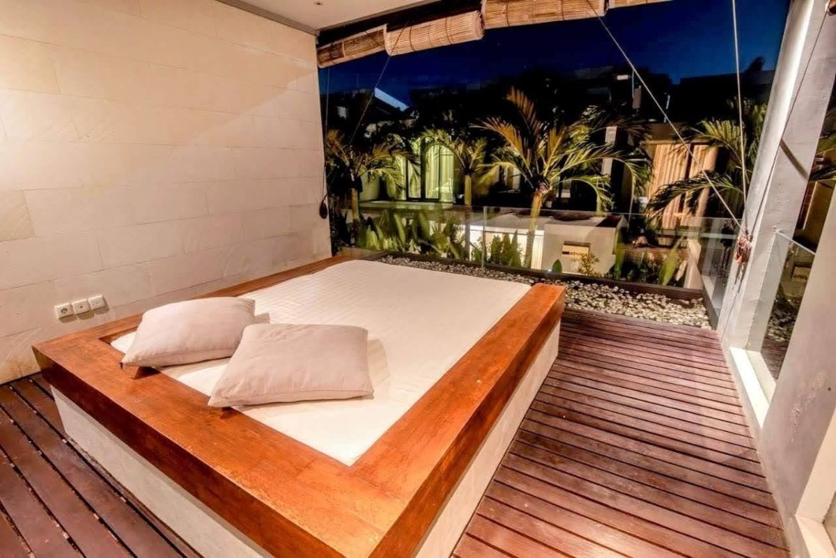 Modern luxury bedroom with king-sized bed, abstract yellow-green wall art, bedside lamps, beige curtains, and woven bench on wooden floor. - Bali Villas