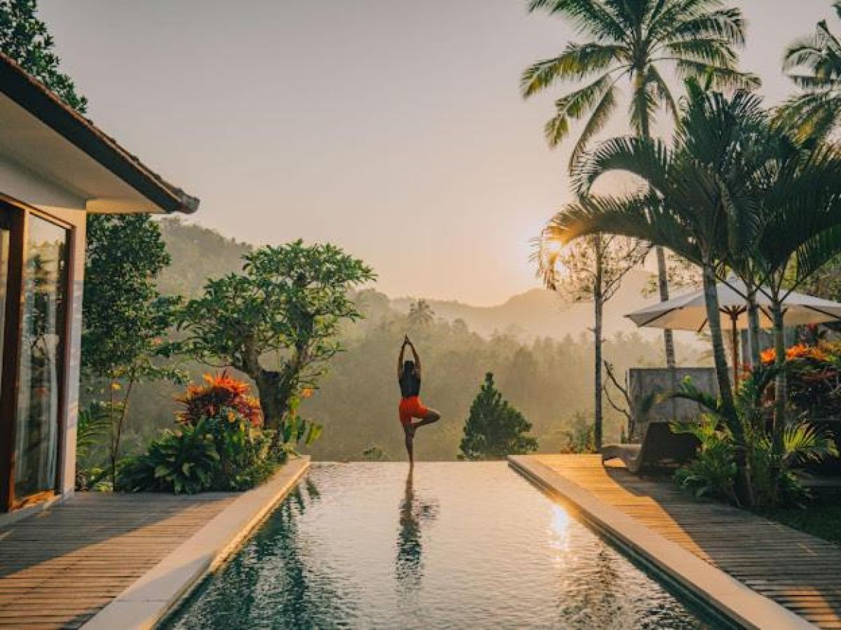 Tropical resort poolside with curved rattan lounge chairs, white umbrella, and lush palm-fringed jungle views. - Bali Villas