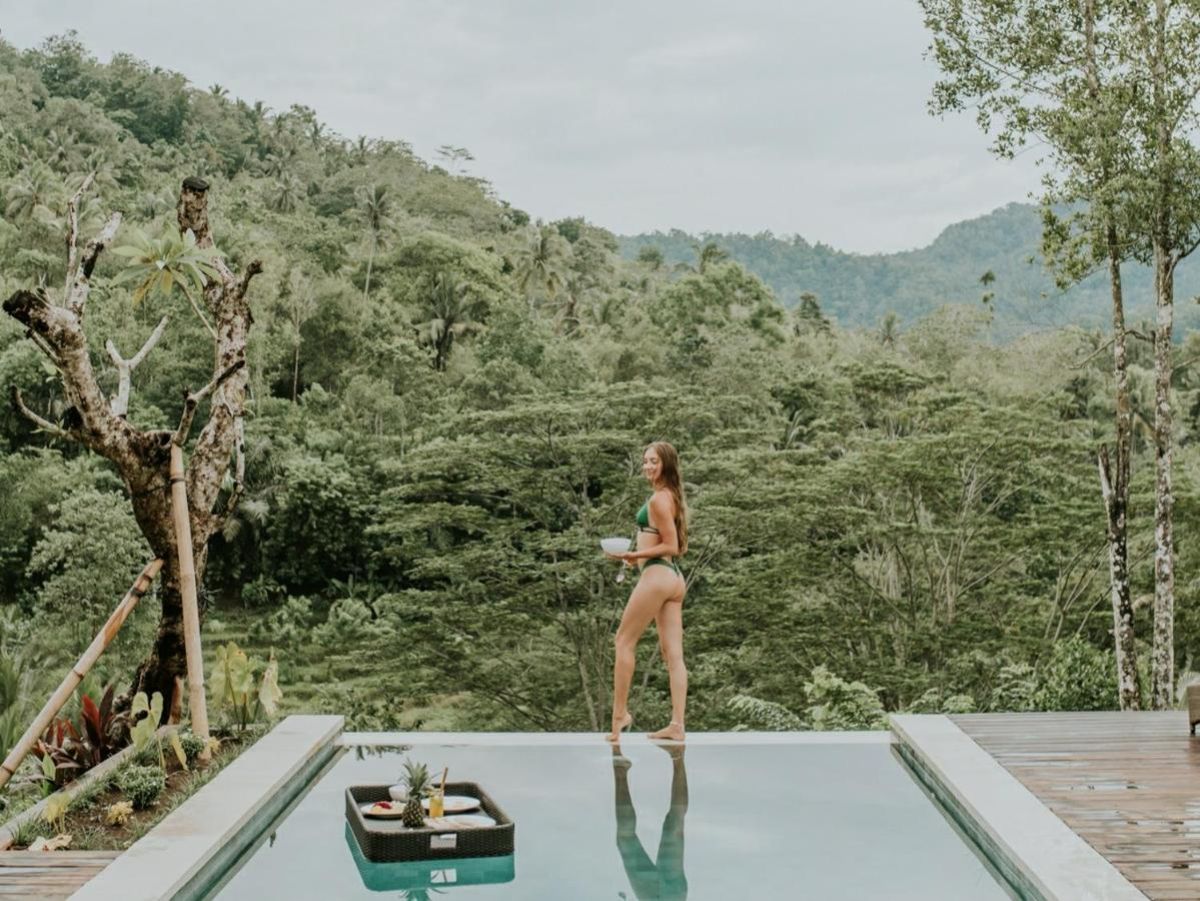 Tropical resort poolside with curved rattan lounge chairs, white umbrella, and lush palm-fringed jungle views. - Bali Villas