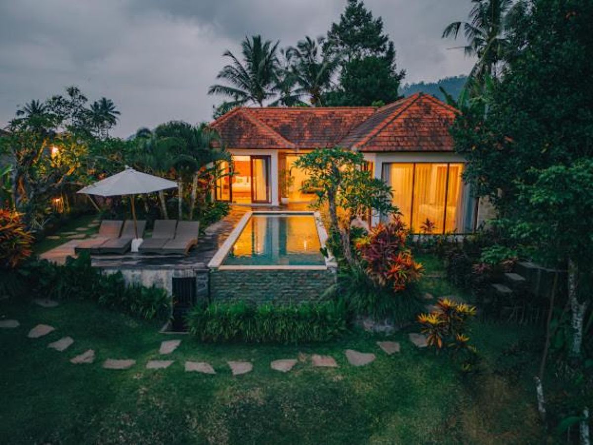 Tropical resort poolside with curved rattan lounge chairs, white umbrella, and lush palm-fringed jungle views. - Bali Villas