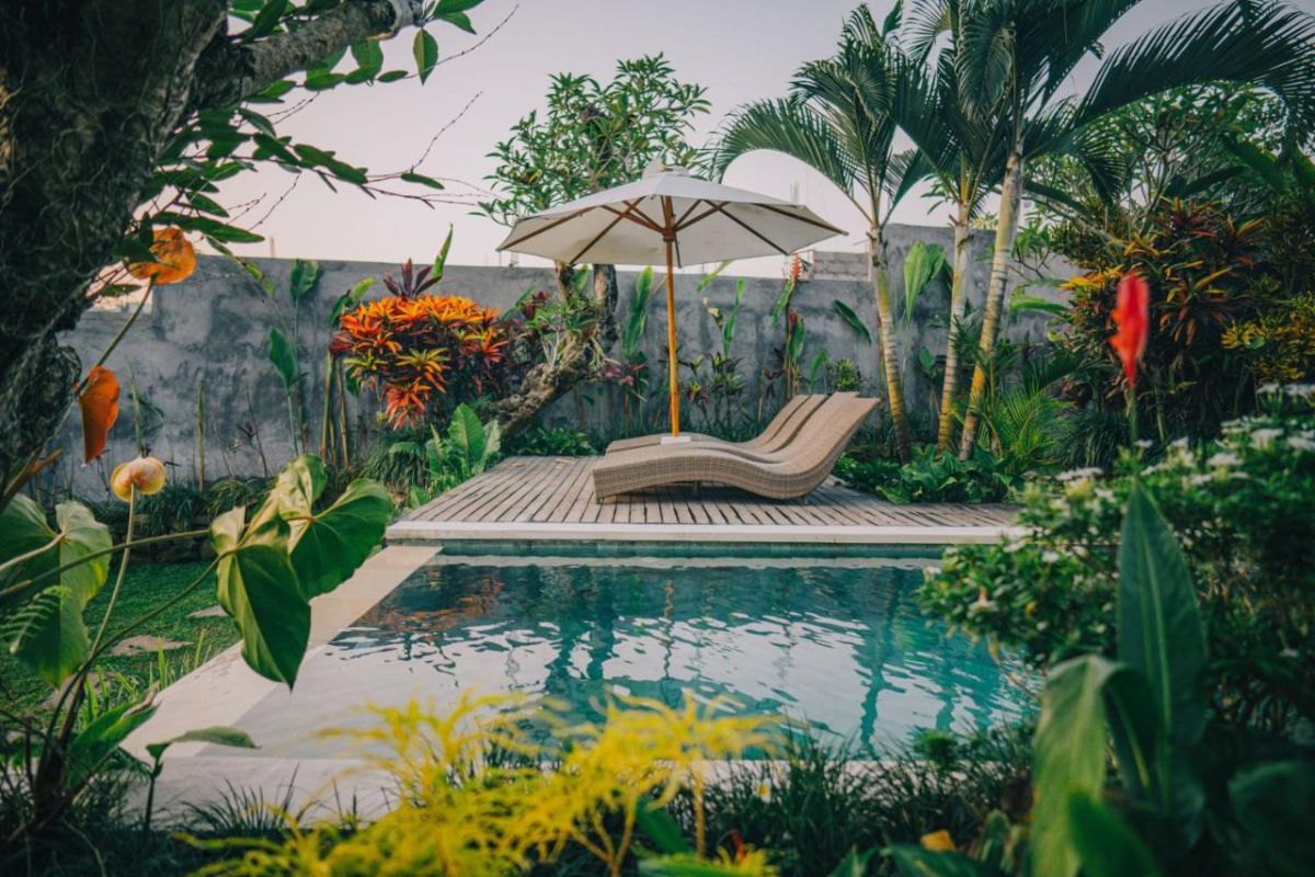 Tropical resort poolside with curved rattan lounge chairs, white umbrella, and lush palm-fringed jungle views. - Bali Villas