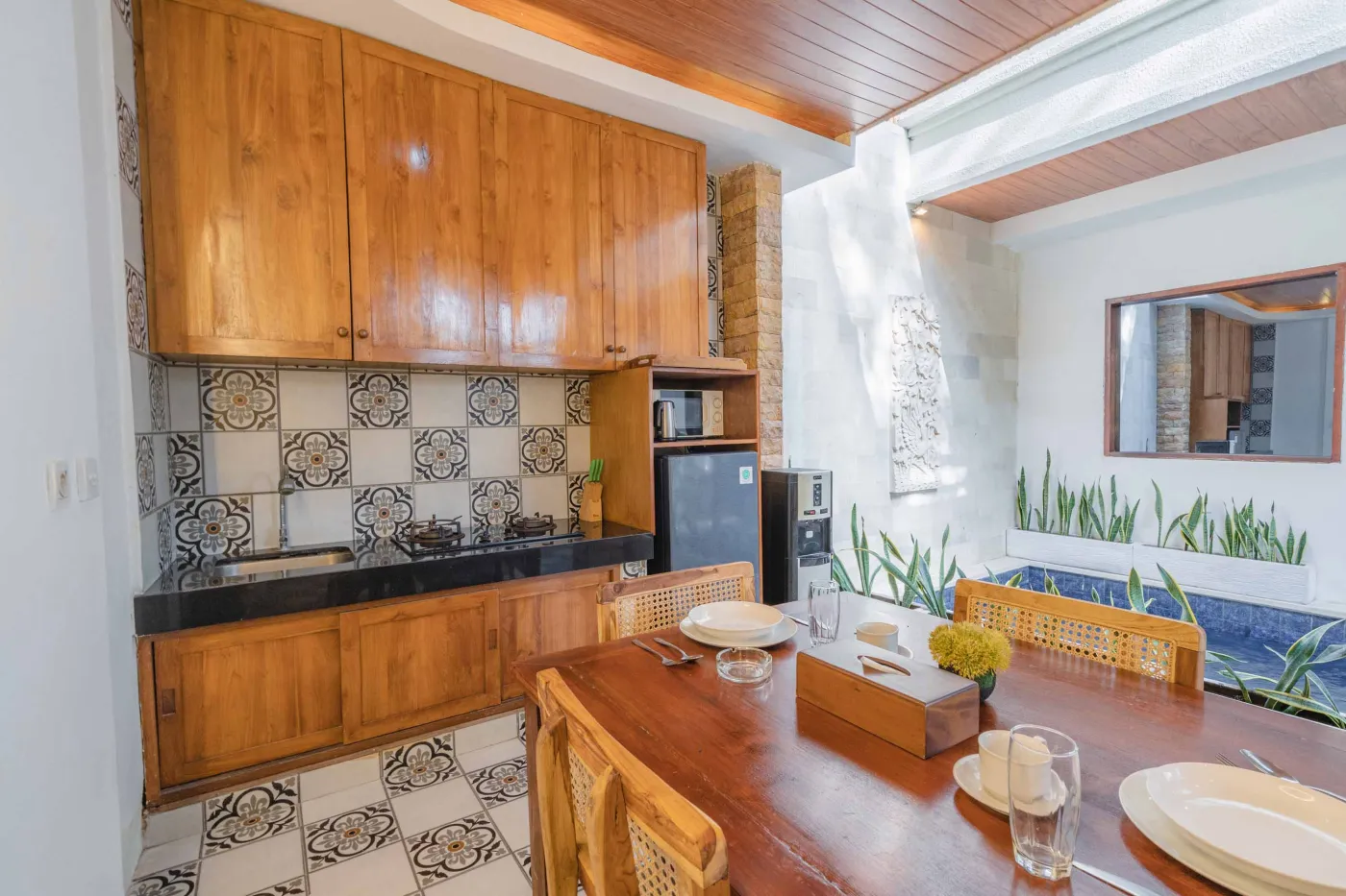 Balinese kitchen with wooden cabinets, checkered tiled backsplash, and dining table overlooking a private pool. - Bali Villas
