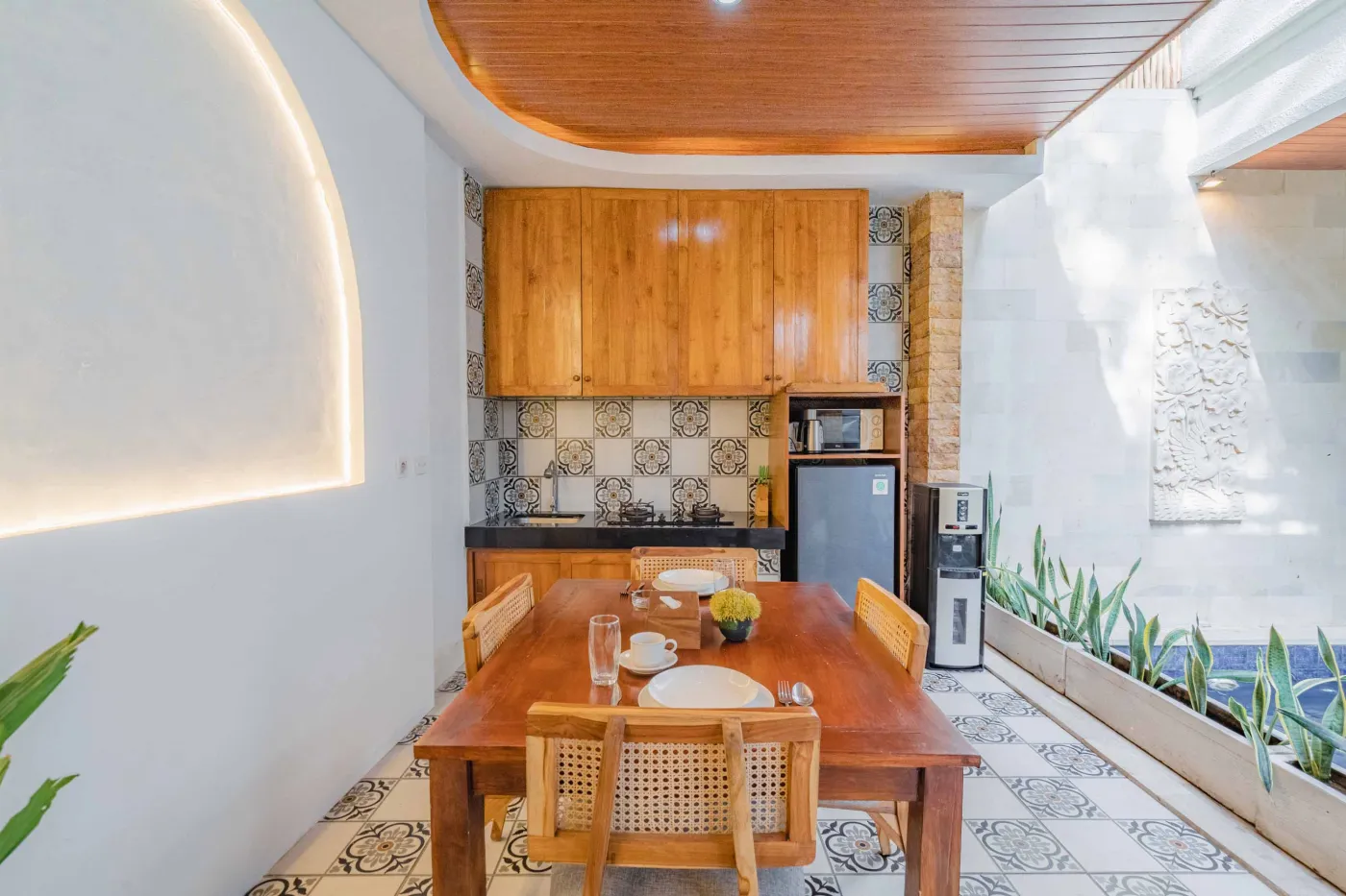 Balinese kitchen with wooden cabinets, checkered tiled backsplash, and dining table overlooking a private pool. - Bali Villas