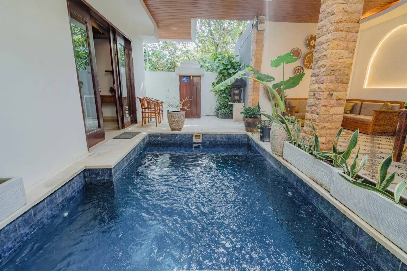 Balinese kitchen with wooden cabinets, checkered tiled backsplash, and dining table overlooking a private pool. - Bali Villas