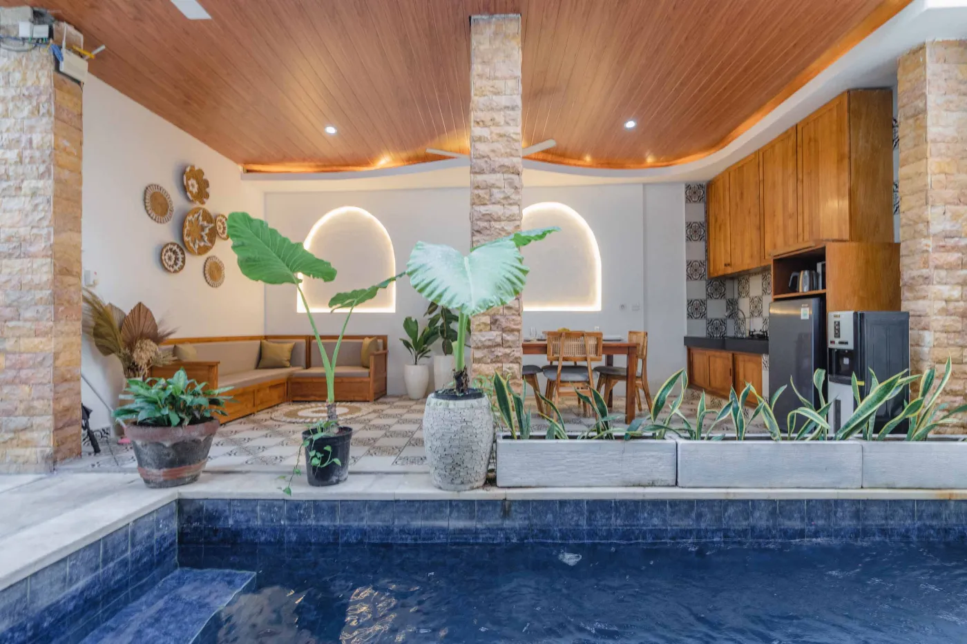 Balinese kitchen with wooden cabinets, checkered tiled backsplash, and dining table overlooking a private pool. - Bali Villas