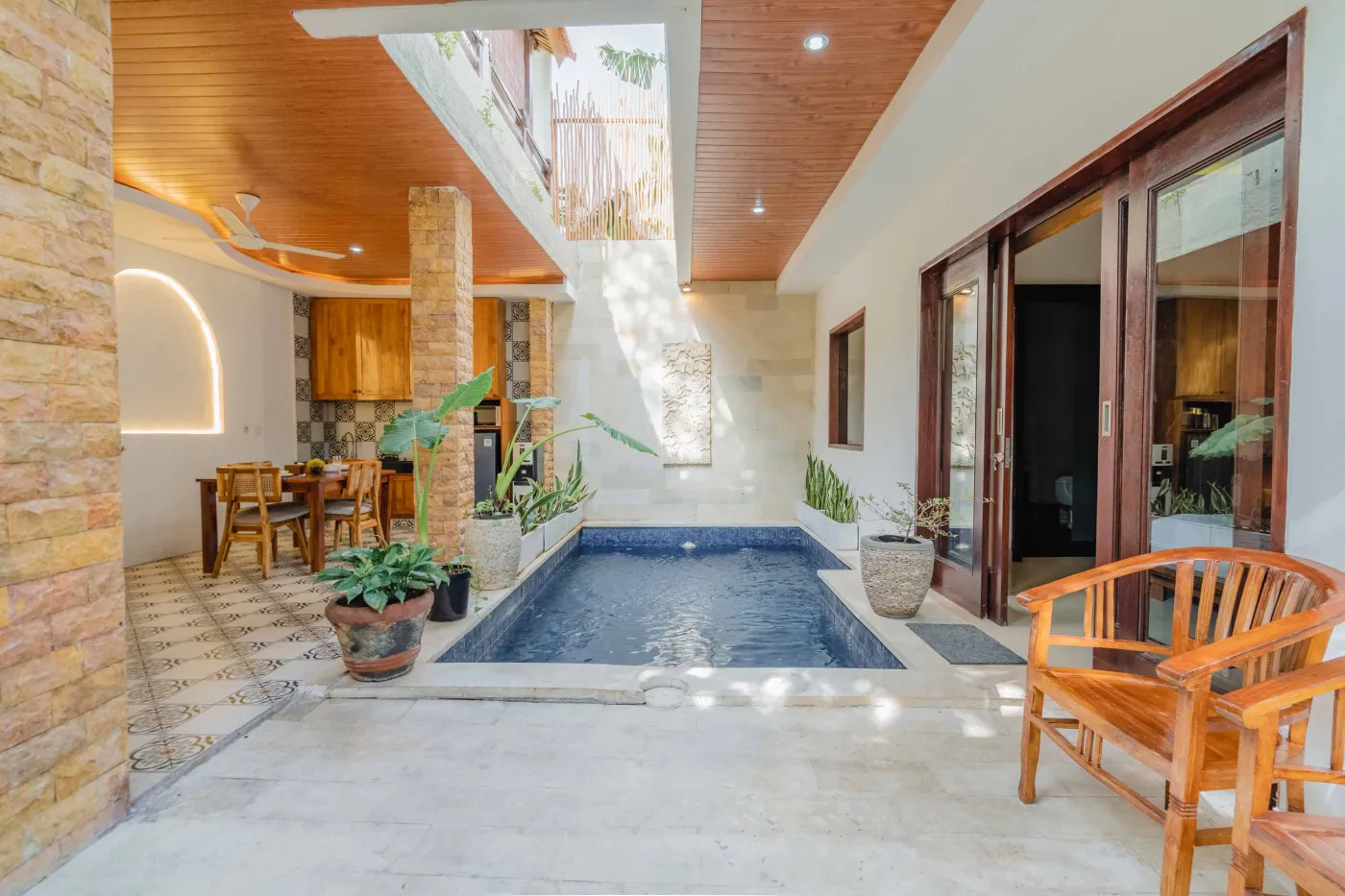 Balinese kitchen with wooden cabinets, checkered tiled backsplash, and dining table overlooking a private pool. - Bali Villas