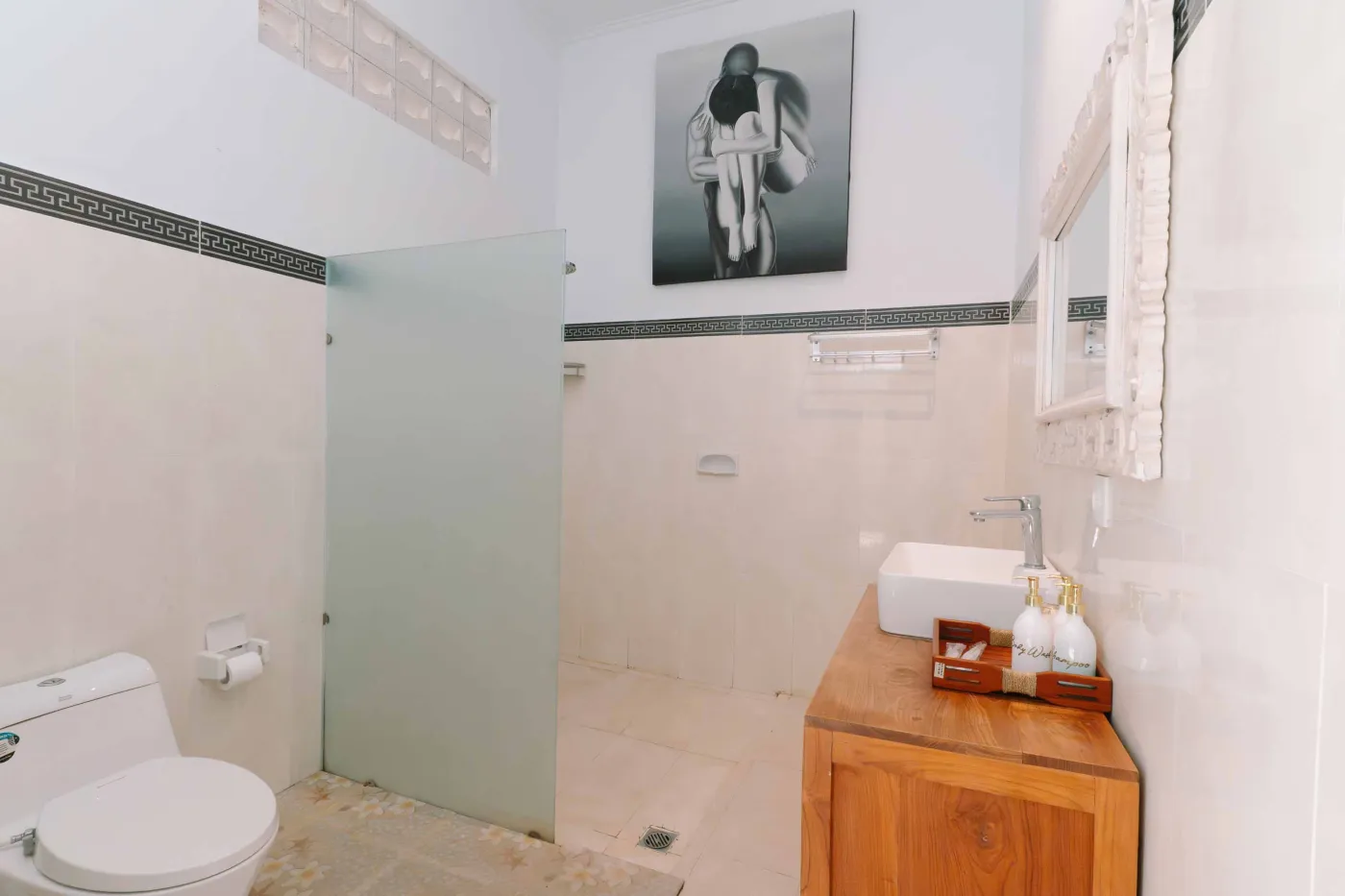 Spacious modern bathroom with white vanity, ornate mirror, toilet, and tropical garden view through frosted glass door. - Bali Villas