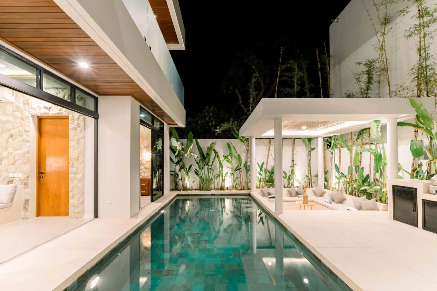 Nighttime view of a modern tropical villa with a glowing turquoise infinity pool, lush bamboo and palm plants, outdoor pergola seating, and barbecue grill. - Bali Villas