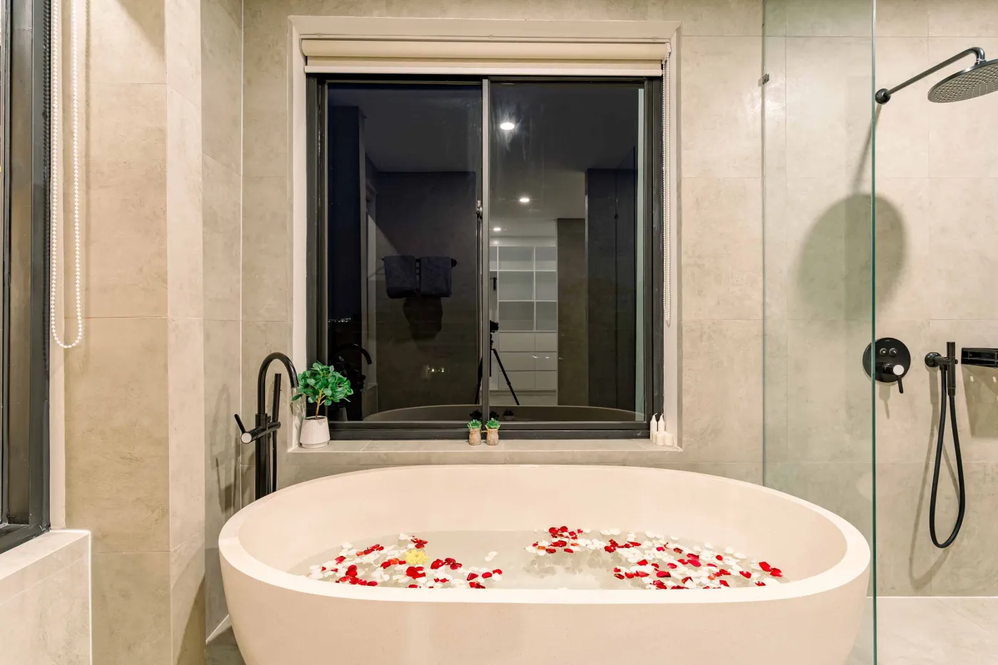 Luxurious modern bathroom featuring a white freestanding bathtub filled with water and red rose petals, adjacent glass shower, large window, and potted plant. - Bali Villas