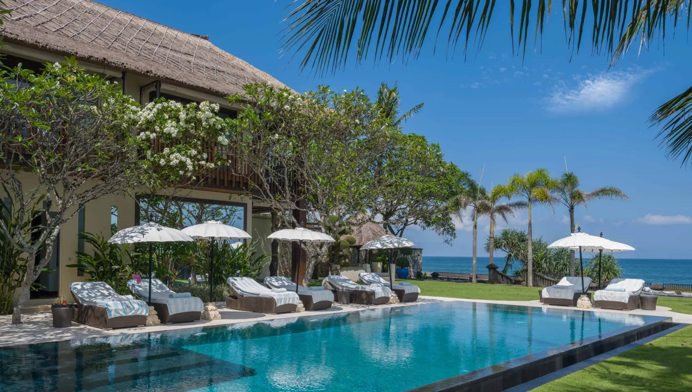 Curved stone terrace with white cushioned lounge overlooking Bali beach, ocean, and tropical gardens. - Bali Villas