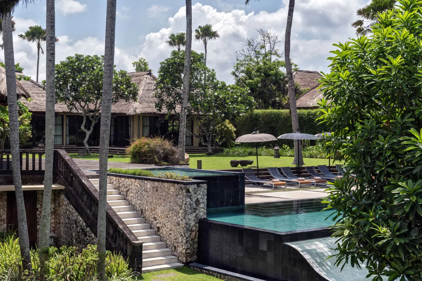 Spacious Balinese pavilion interior with thatched roof, white modular sofas, woven accents, and lush garden view through open wooden frames. - Bali Villas