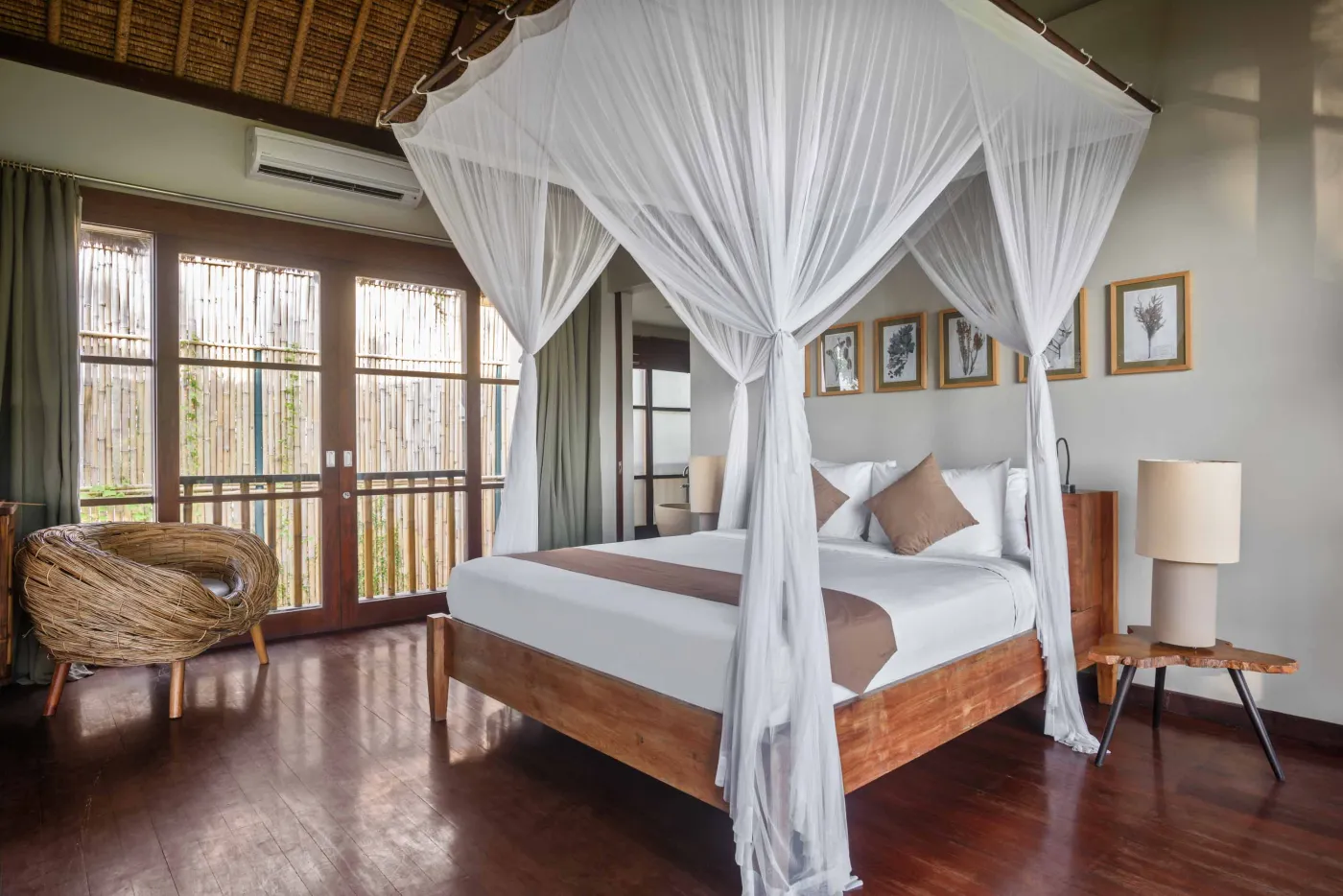 Spacious Balinese pavilion interior with thatched roof, white modular sofas, woven accents, and lush garden view through open wooden frames. - Bali Villas