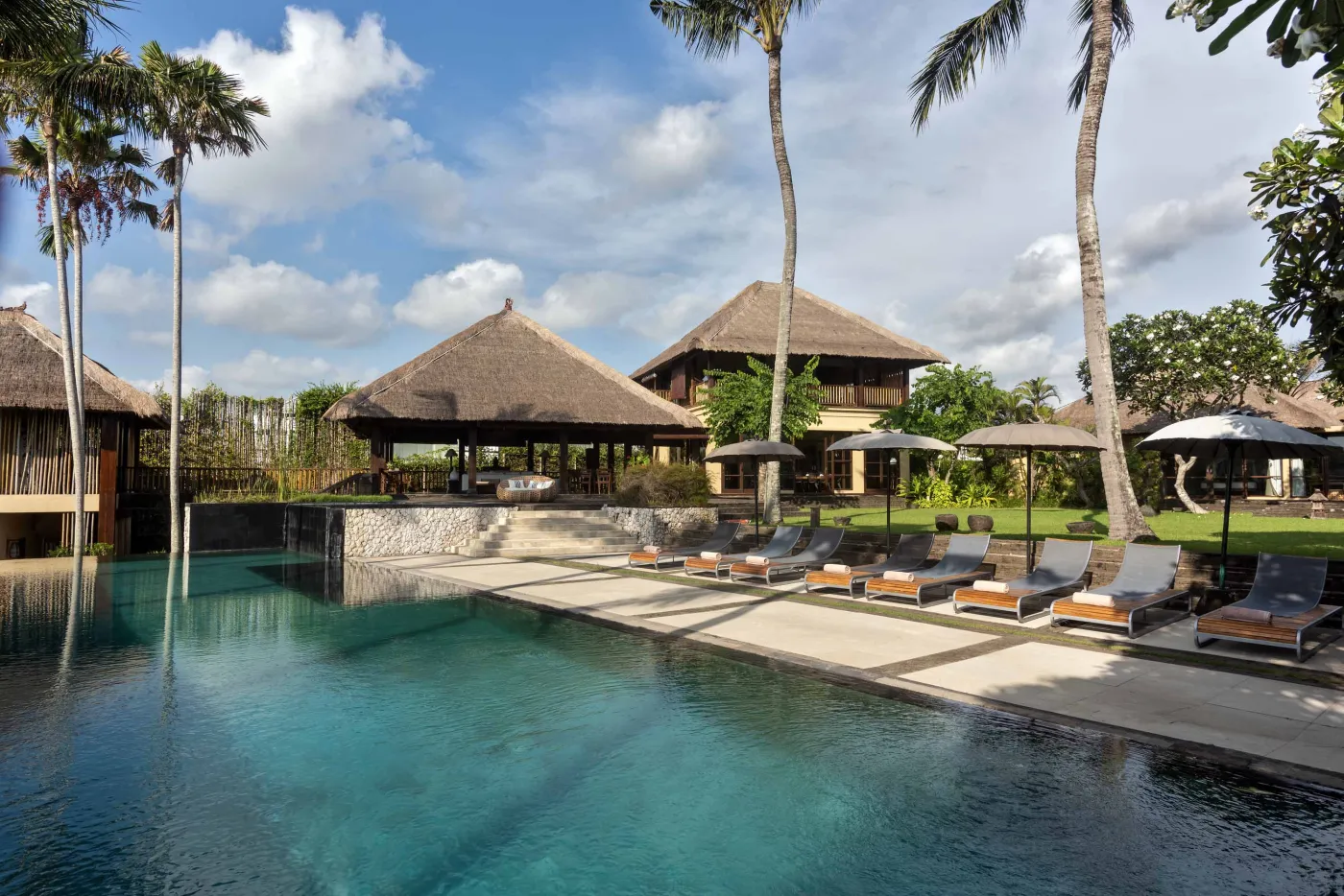Spacious Balinese pavilion interior with thatched roof, white modular sofas, woven accents, and lush garden view through open wooden frames. - Bali Villas