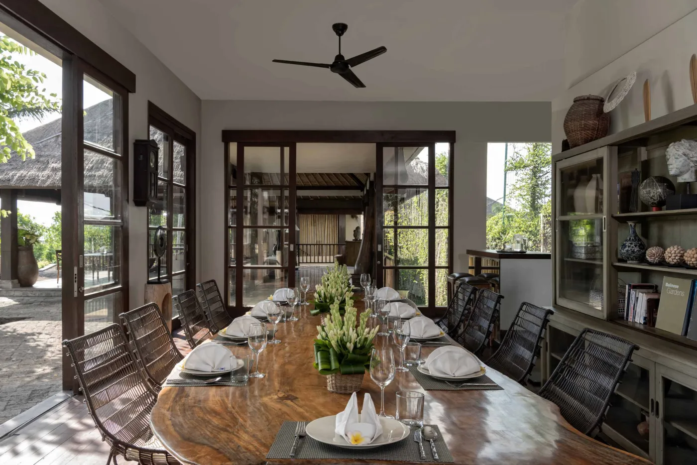 Spacious Balinese pavilion interior with thatched roof, white modular sofas, woven accents, and lush garden view through open wooden frames. - Bali Villas