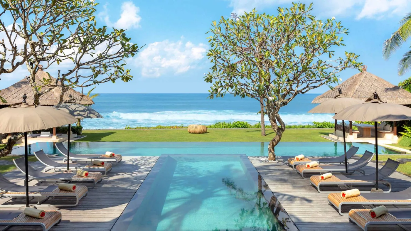 Luxury infinity pool at Bali beach resort, with loungers, umbrellas, tropical trees, and ocean view. - Bali Villas