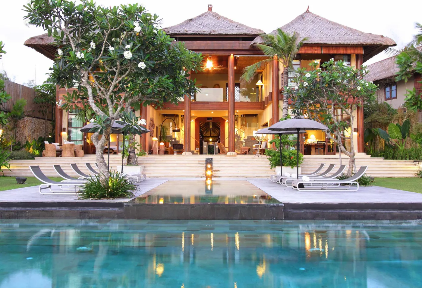 Luxurious Balinese villa with infinity pool, tropical frangipani trees, and outdoor loungers at sunset. - Bali Villas